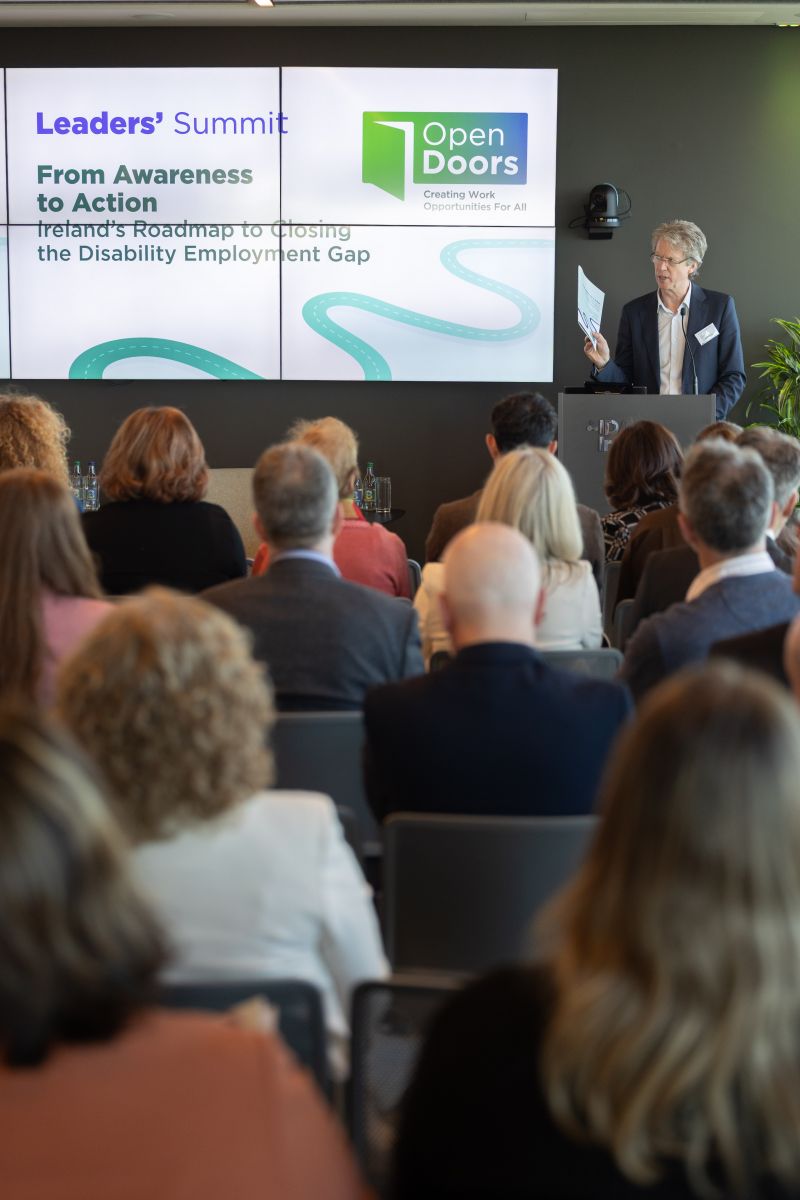 Large seated audience watching a presentation by a blonde woman wearing a blue dress speaking at podium. Screen with Open Doors logo reading Leaders' Summit